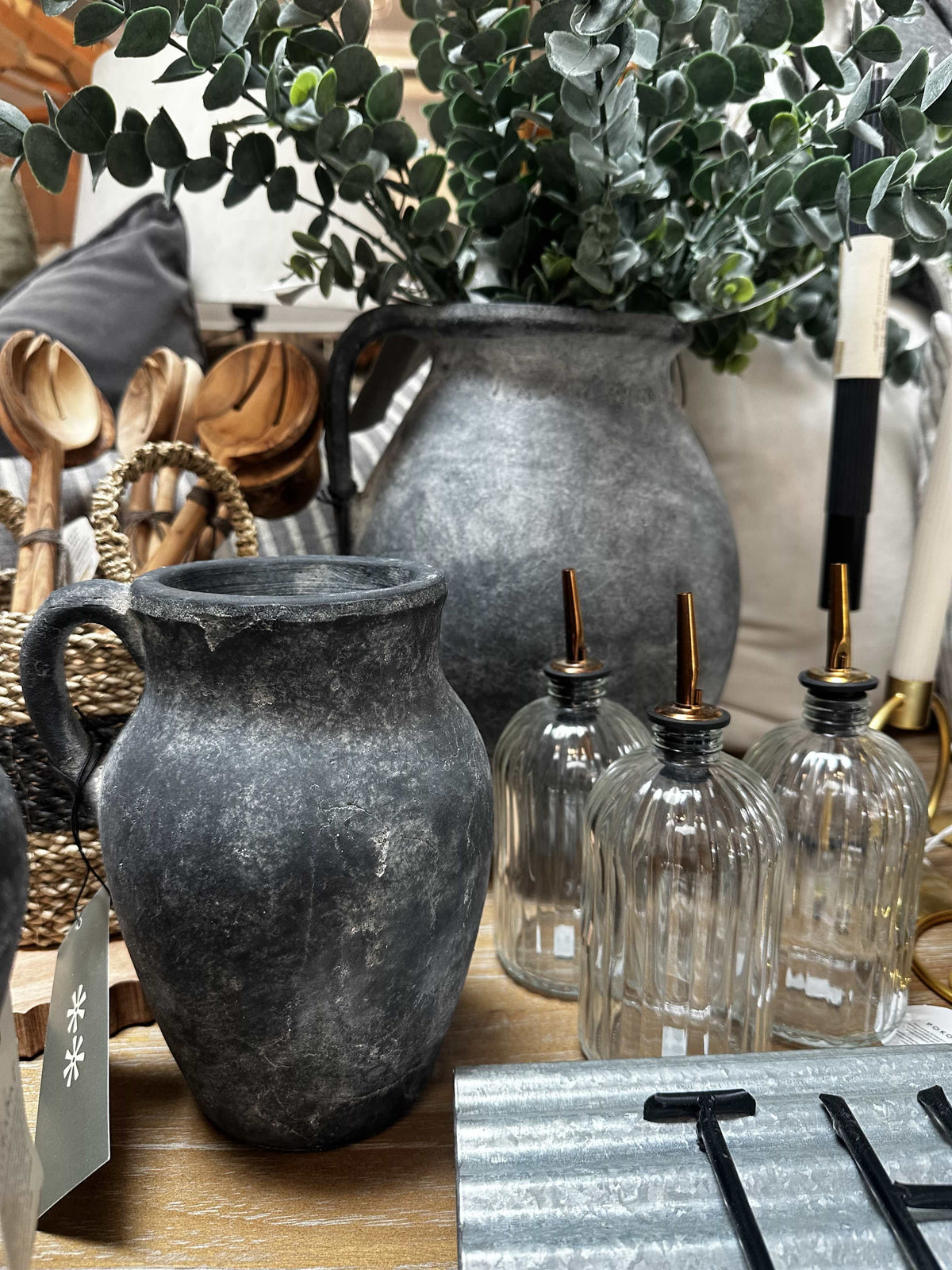 A rustic dark gray stoneware pitcher with a handle sits on a wooden table alongside three ribbed clear glass bottles with metal pourers, with a larger matching stoneware vase and greenery in the background.